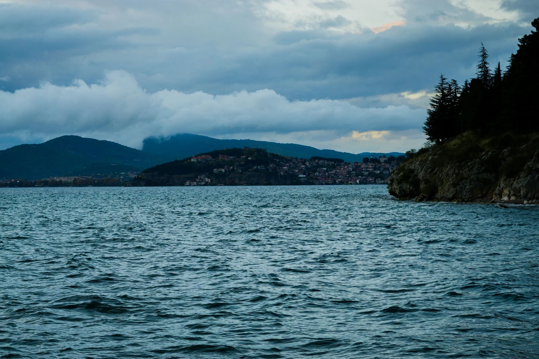 Lake Ohrid with a town and mountains beneath a cloudy sky.